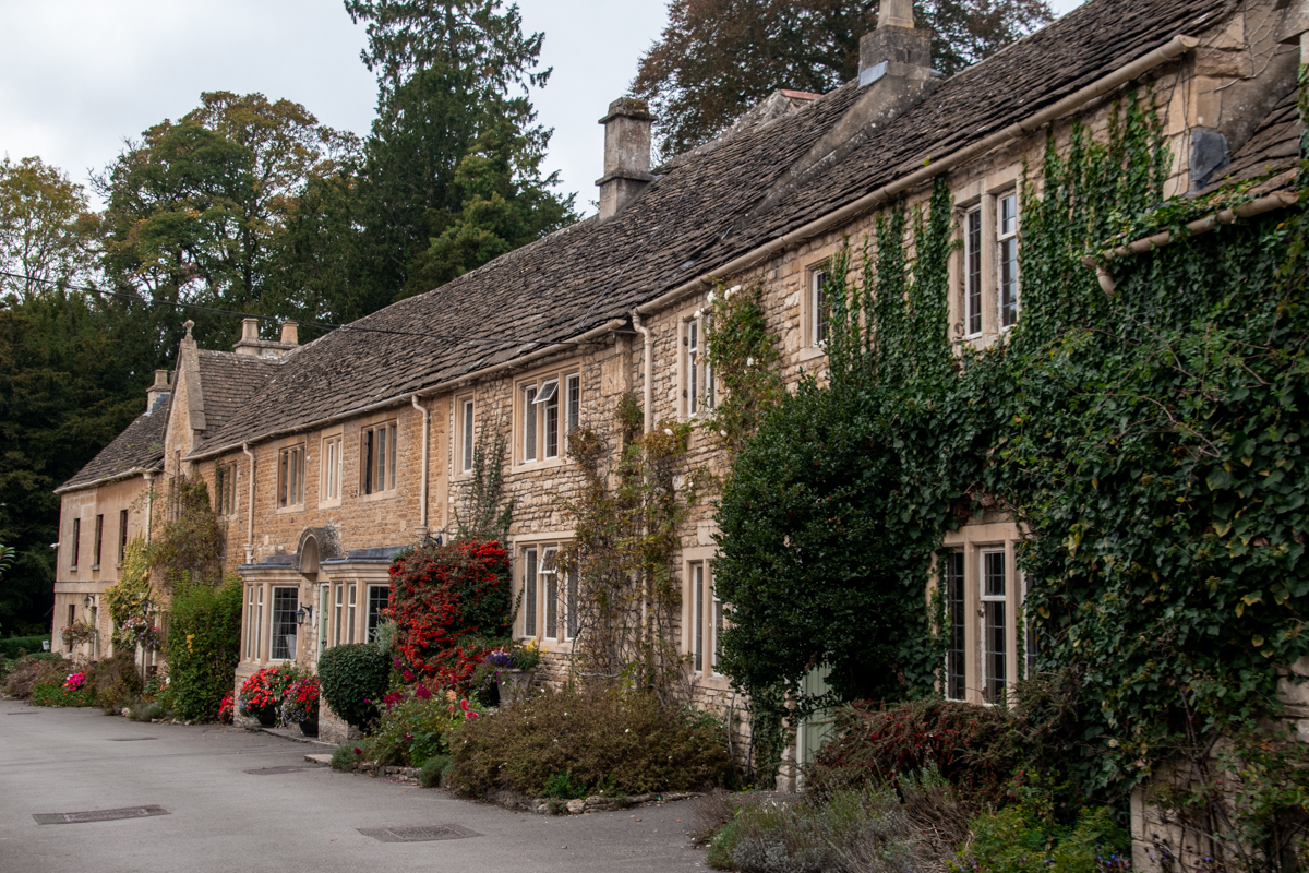 Row of historic Cotswold stone cottages covered in ivy and flowers on West Street in Castle Combe.