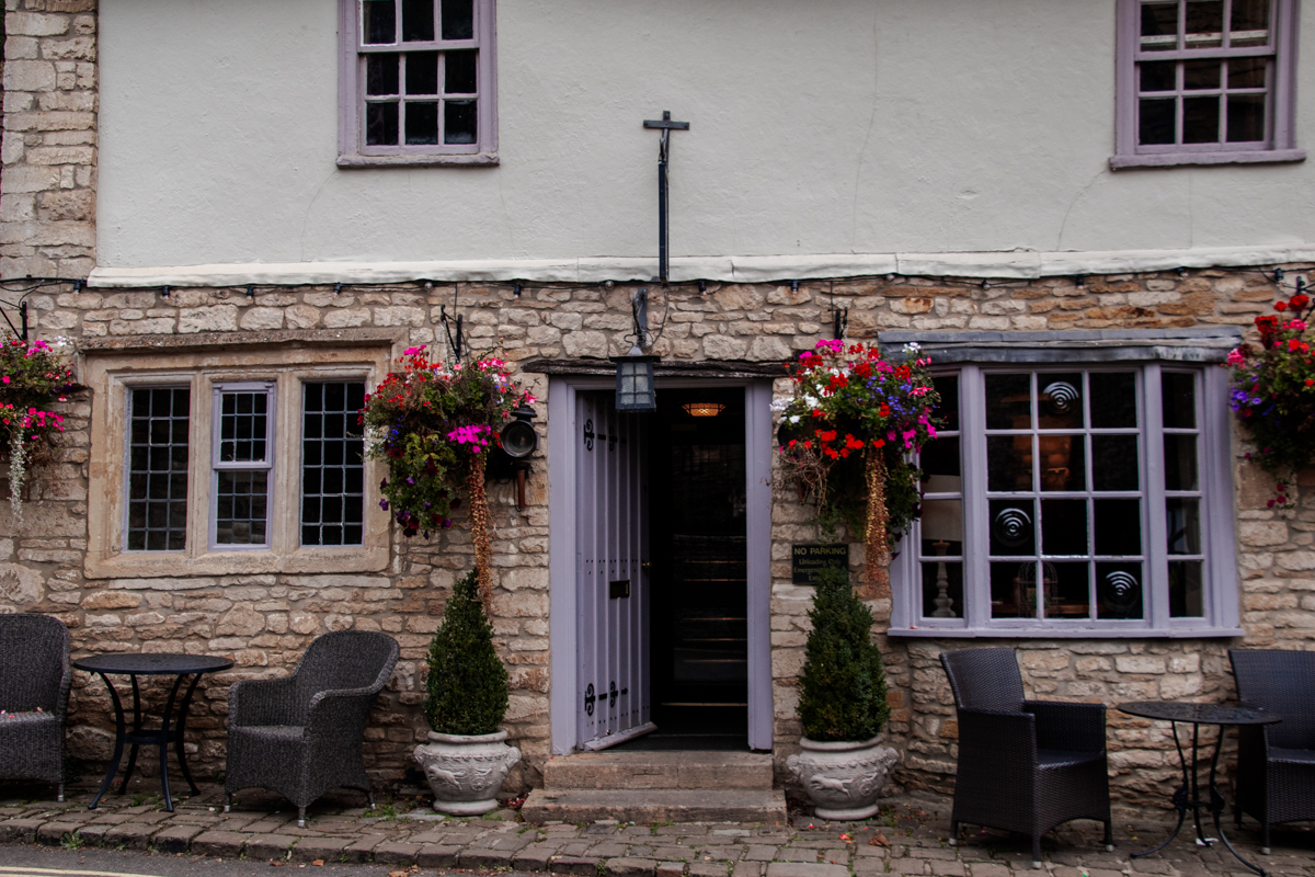Stone facade of The Castle Inn in Castle Combe with purple-trimmed windows, hanging flower baskets and outdoor seating.